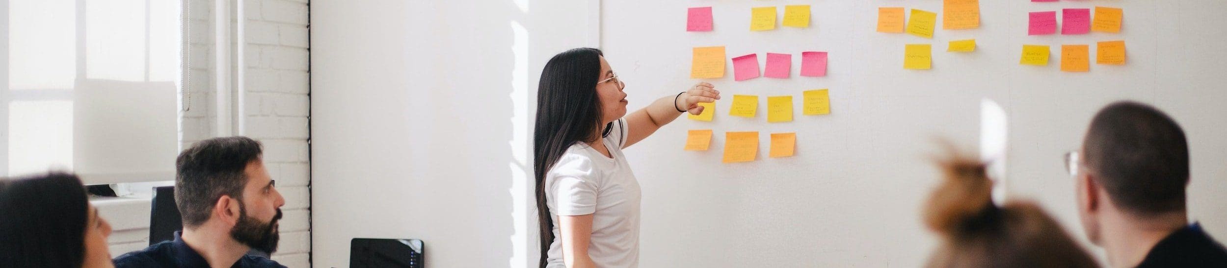 a young woman stood near a white board with sticky notes all over it, she is explaining something to the rest of the people in the room.