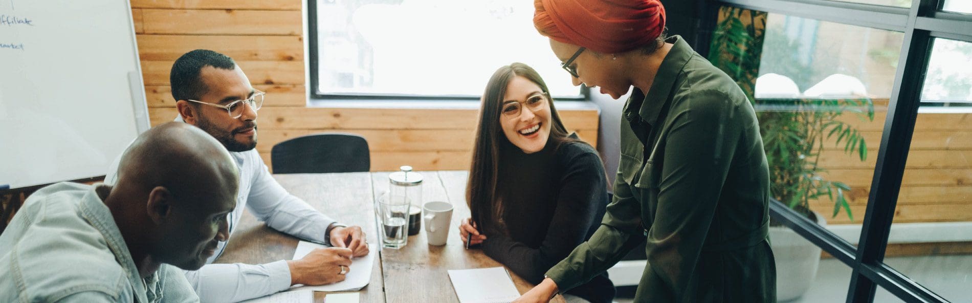 A group of diverse business people sat round a table, all of them are smiling and they are looking at paperwork. The lead female is stood up.