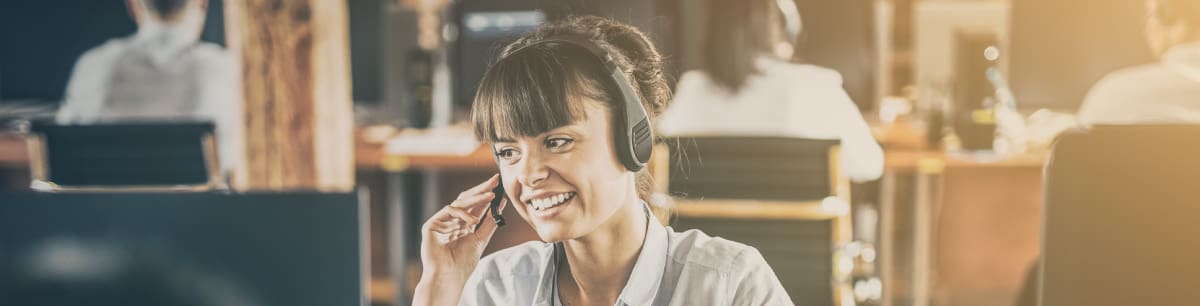 Woman in an office smiles at her screen, talking on her headset and holding the mouthpiece.