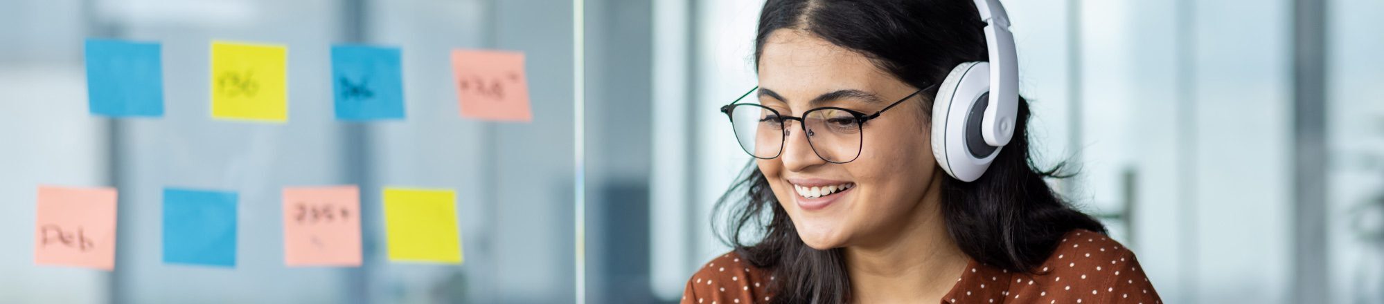 Smiling woman wearing glasses and white earphones, working on a laptop in an office setting.