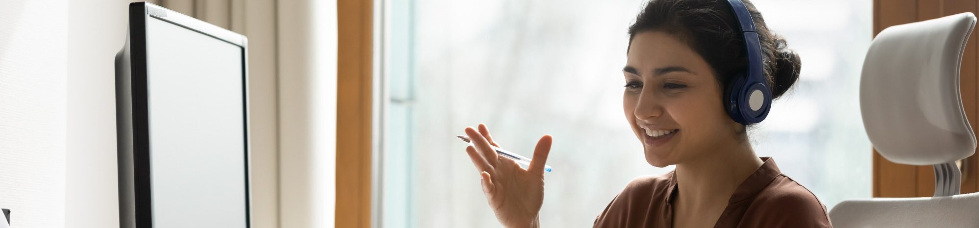 Smiling woman gesturing wearing blue earphones, working on a laptop, bright window behind her.