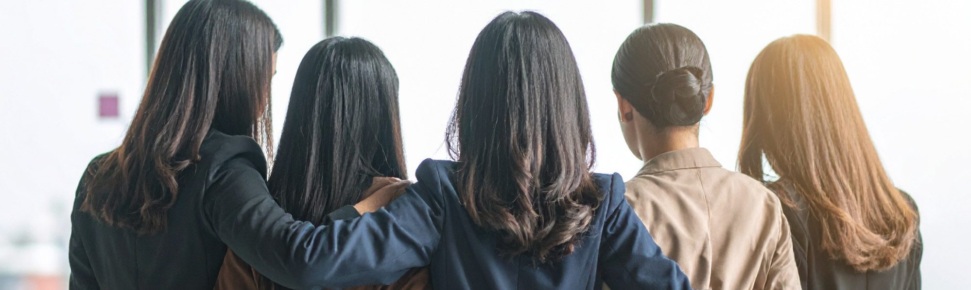 Five business woman stood with their backs to the camera, arms around each others backs - modern office setting.