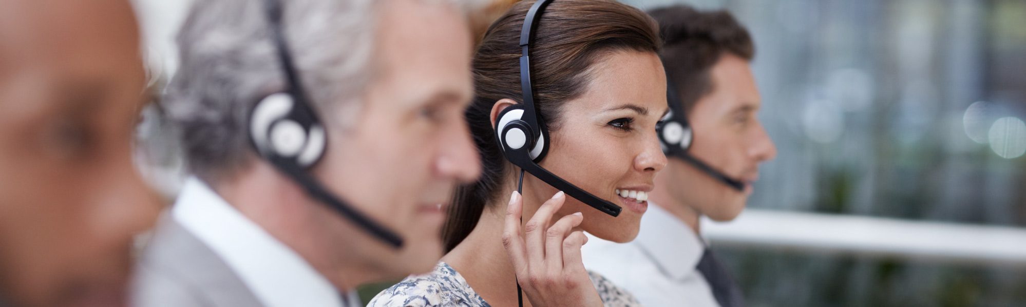 A row of business people all sat with headsets on, the focus is on one woman talking and smiling at the screen.