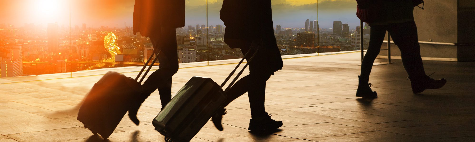 Three people all dragging suitcases in an airport, they are in silhouette with a sunset background over a city behind.