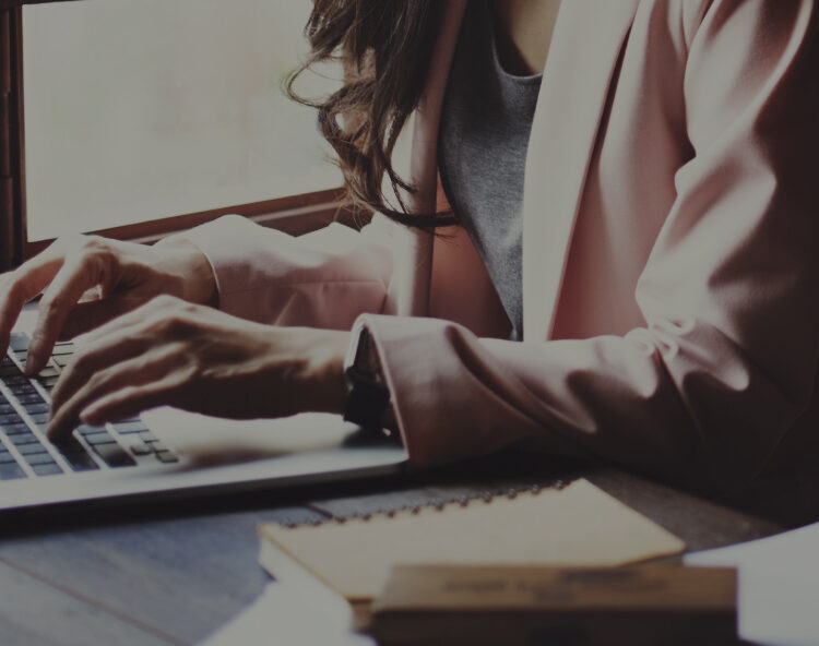 Close-up of a person typing on a laptop beside notebooks on a wooden desk.