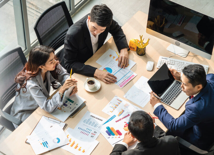 Four people review printed charts and graphs around a desk with laptops and office items.
