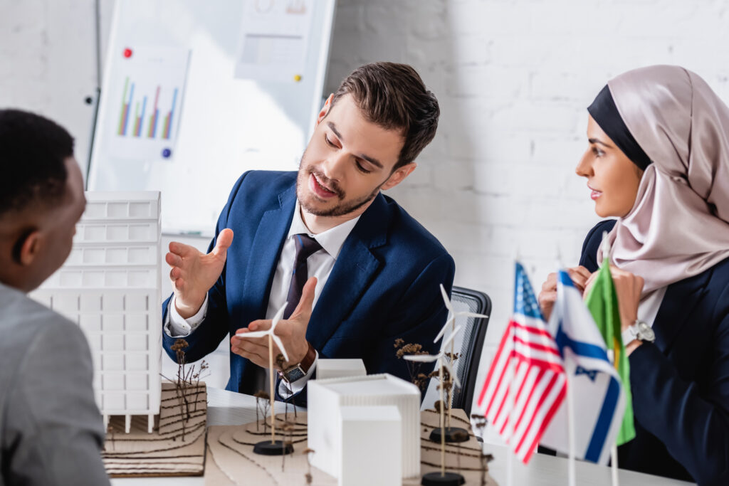 Three people sit at a table discussing a scale building model and pointing at design details.