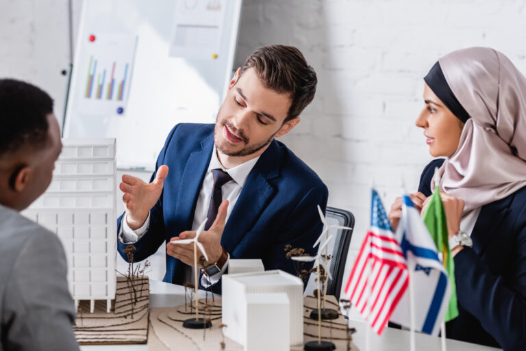 Three people sit at a table discussing a scale building model and pointing at design details.