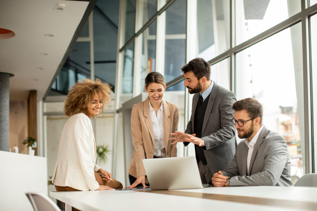 Four people stand at a table discussing work and looking at a laptop together.