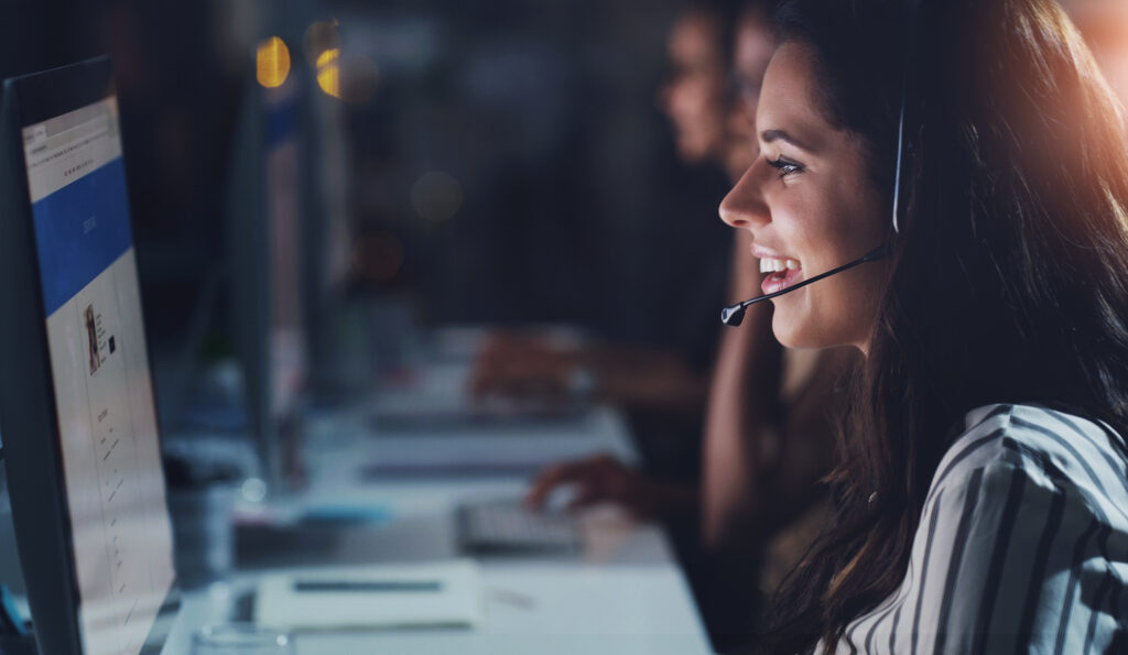 Person wearing a headset uses a computer at a desk, with others working in the background.