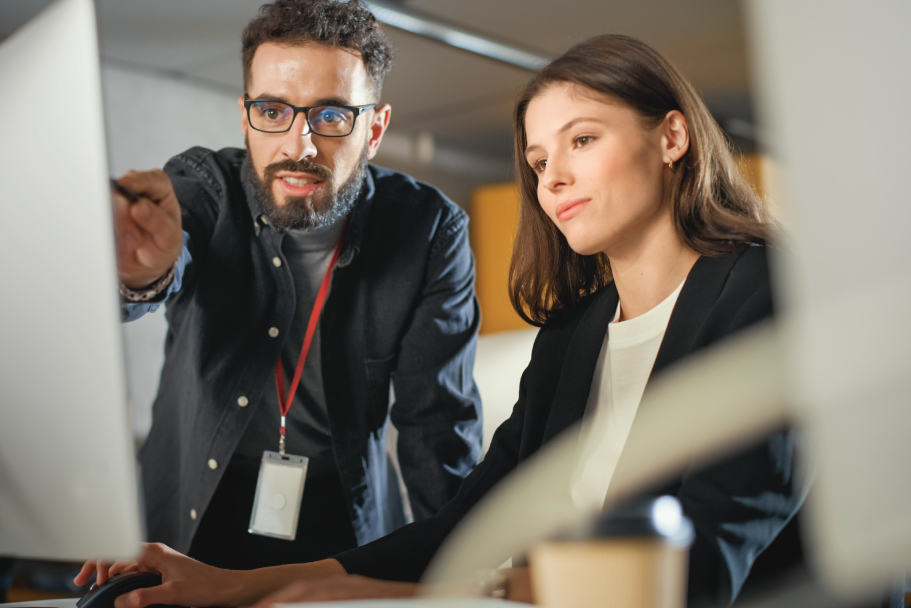 Two people look at a computer screen, one pointing while discussing work.