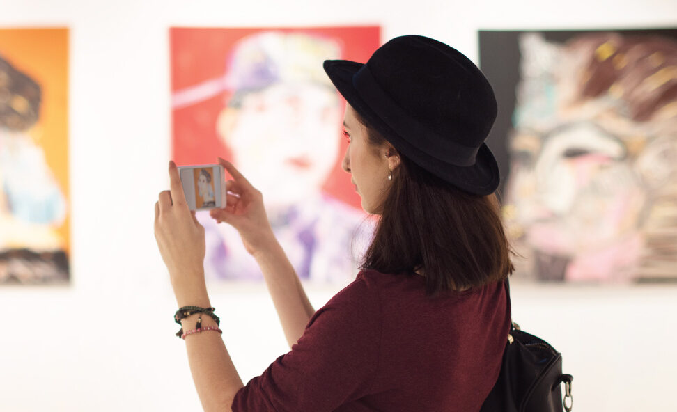 Person wearing a hat takes a photo of paintings displayed on a gallery wall.