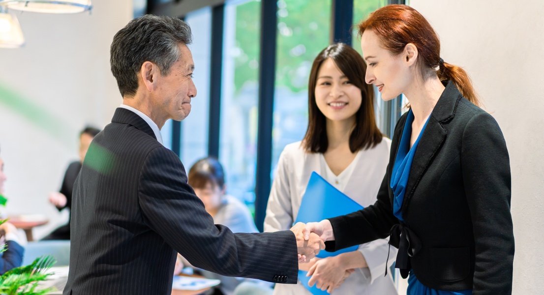 Two professionals shaking hands in an office while a colleague stands behind holding a folder.