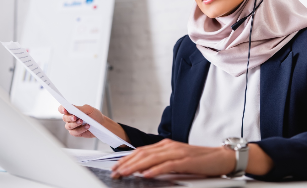 Person wearing a headset reviewing printed documents beside a laptop on a desk.