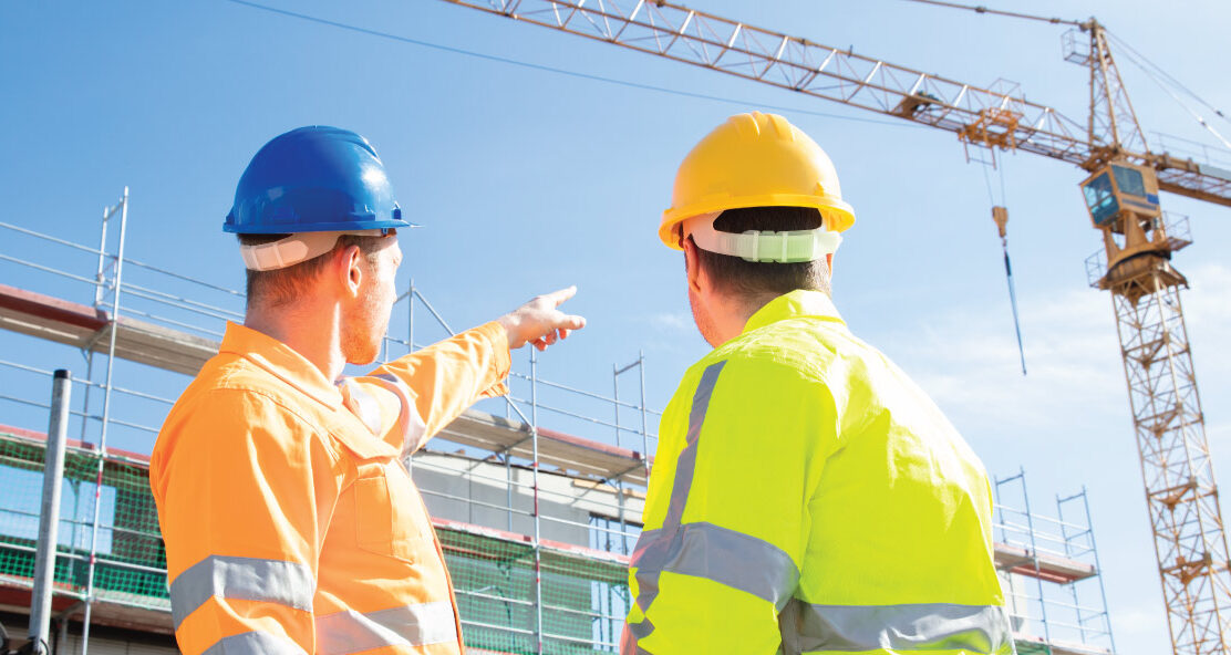 Two people in safety helmets stand on a construction site, one pointing towards a crane.
