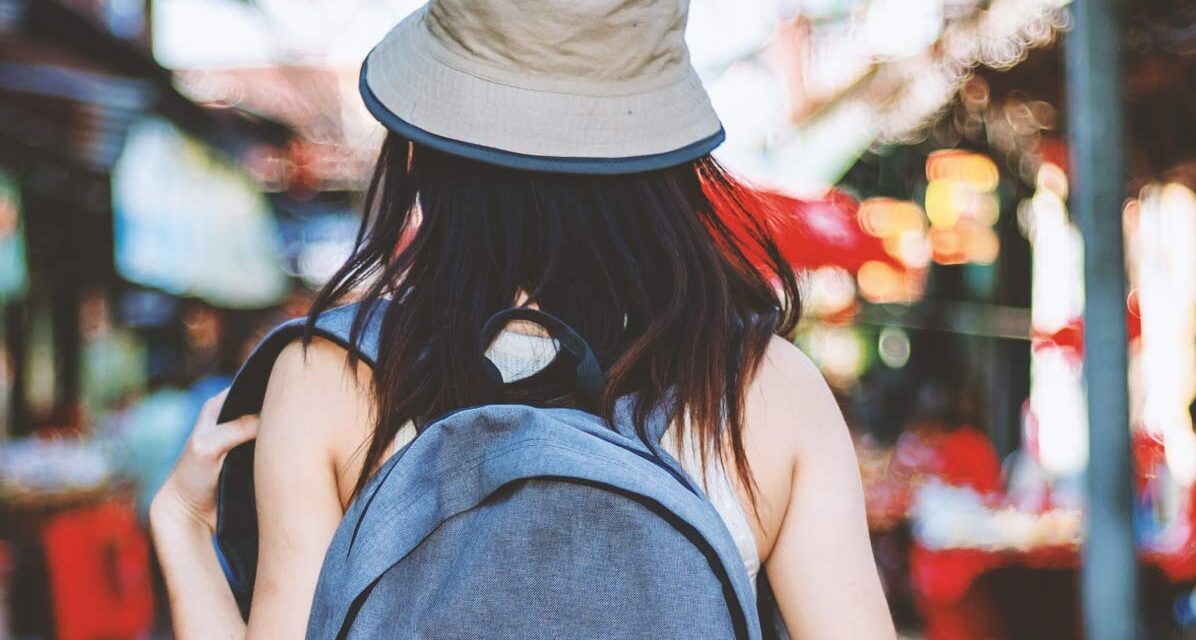 Person wearing a hat and backpack walks through a busy street market with stalls.