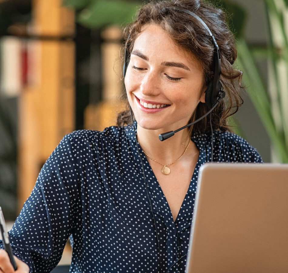 Person wearing a headset uses a laptop and writes notes while seated at a desk.