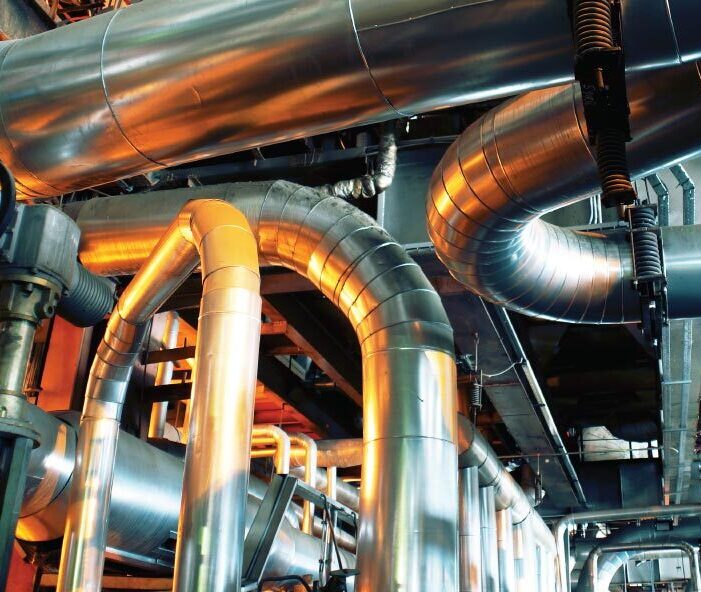 Network of large metal pipes and valves inside an industrial facility with overhead structures.