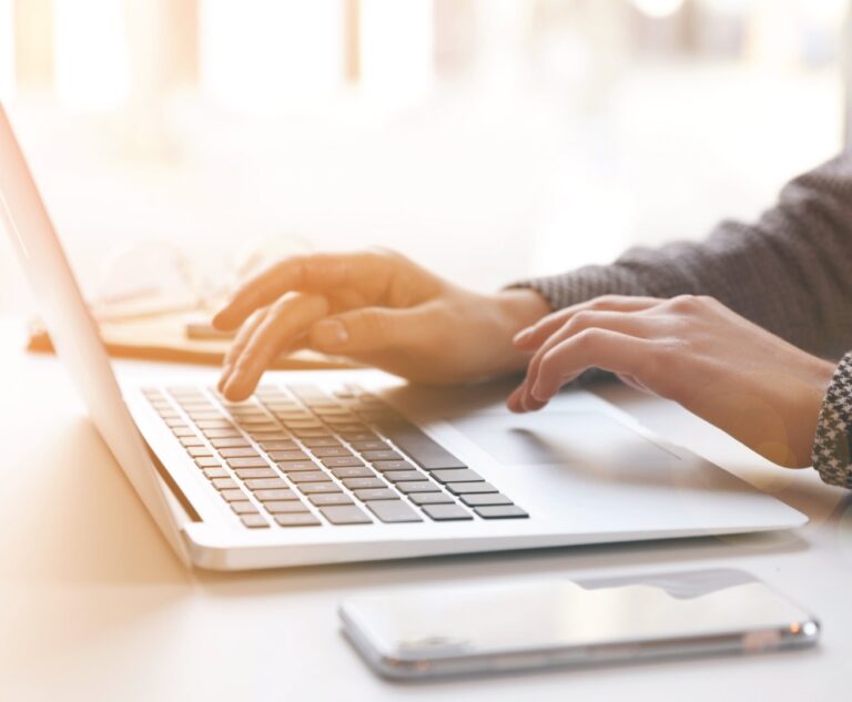 Close‑up of hands typing on a laptop with a smartphone resting on a desk.