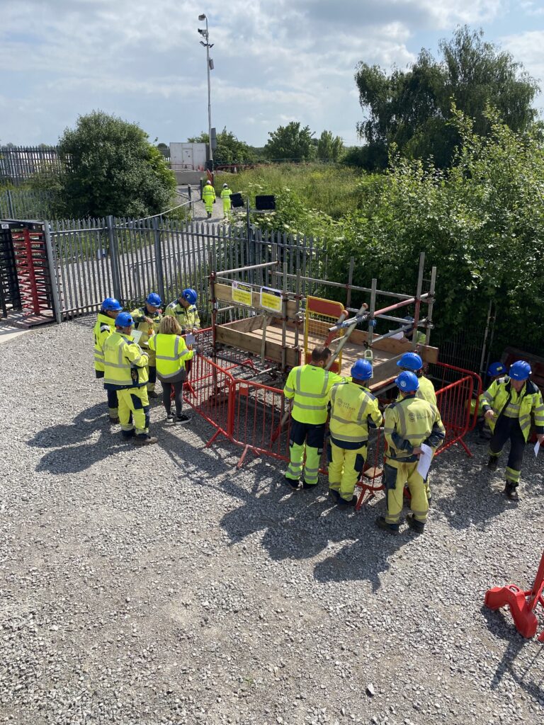 Group of workers in safety helmets and high-visibility clothing gathered around equipment on site.