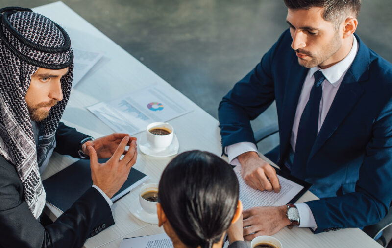 Three people seated at a table review documents and discuss work over coffee.