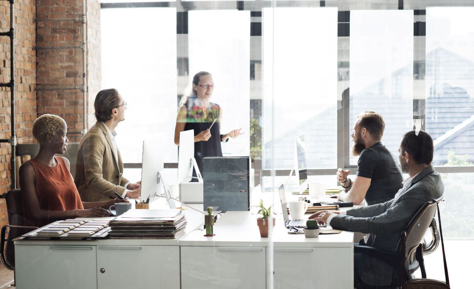 One person presents to a group seated at desks with laptops in a bright office.
