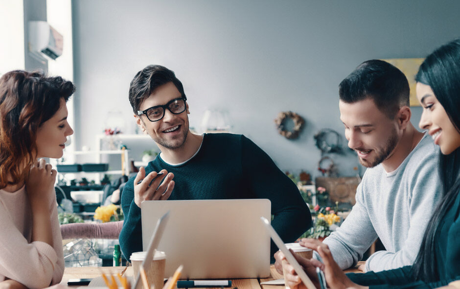 Four people sit around a table using a laptop and tablet while discussing work together.