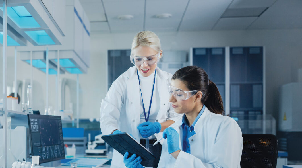 Two people in lab coats and safety glasses review data on a tablet in a laboratory.