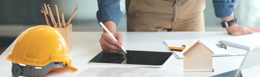 Close-up of a hand using a stylus on a tablet beside a safety helmet and pencils.