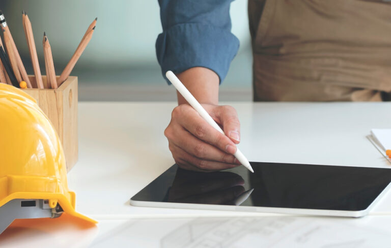 Close-up of a hand using a stylus on a tablet beside a safety helmet and pencils.