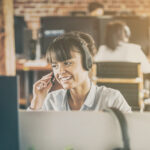 Person wearing a headset speaks while working at a desk in an open office environment.