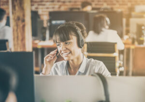 Person wearing a headset speaks while working at a desk in an open office environment.