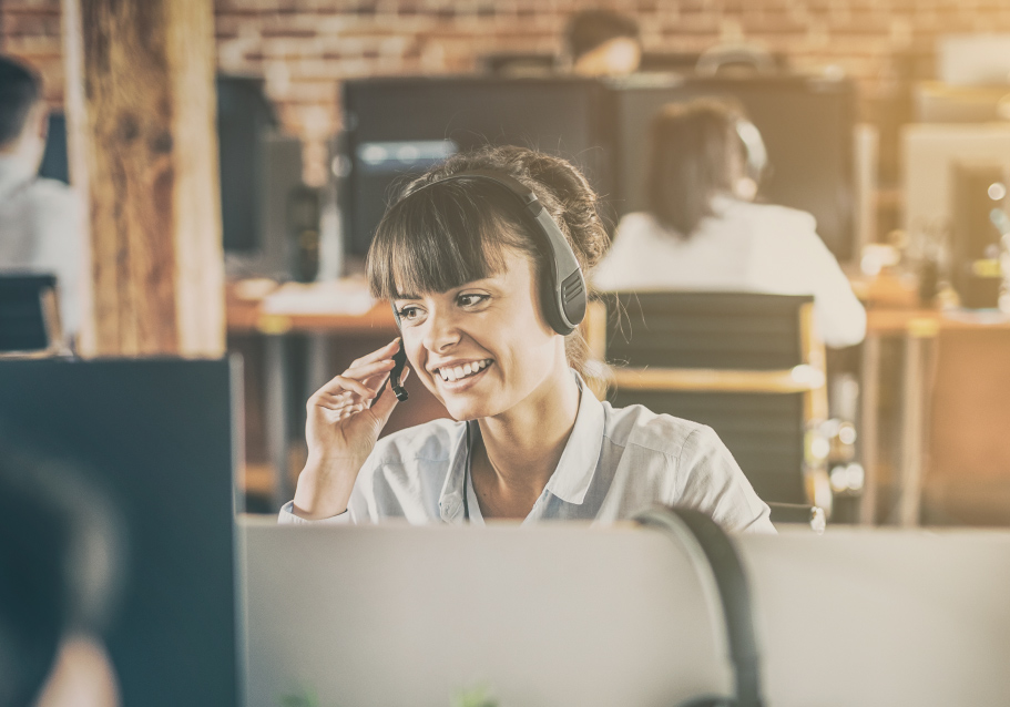 Person wearing a headset speaks while working at a desk in an open office environment.