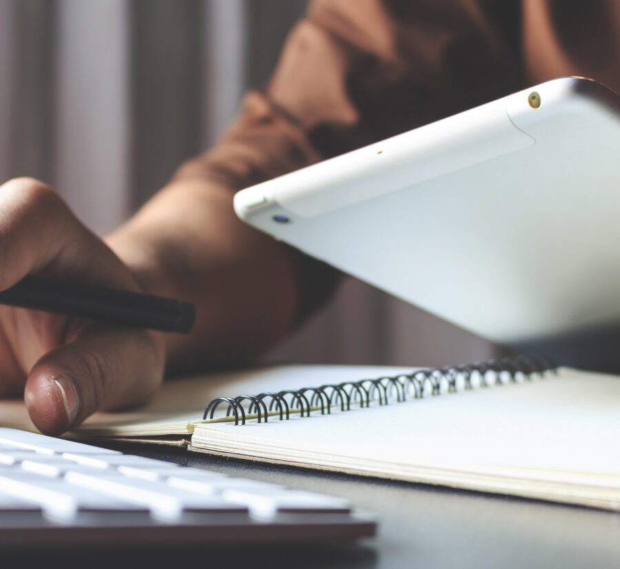 Close-up of a person writing in a notebook beside a tablet and keyboard at a desk.