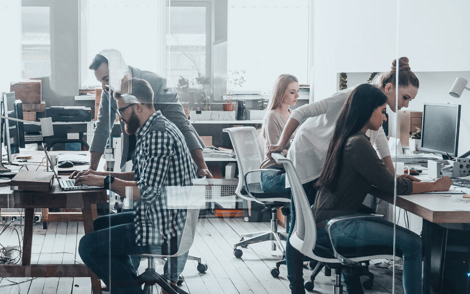 Several people sit and stand at desks using computers in a bright open office space.