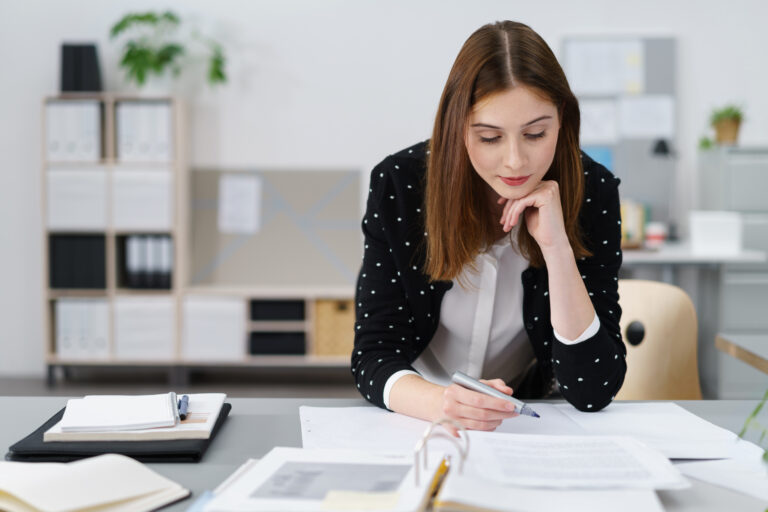 Person seated at a desk reading documents and holding a pen, with files and folders nearby.