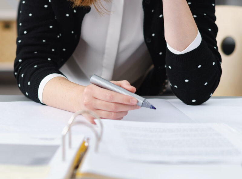 Close-up of a person holding a pen and writing on printed documents at a desk.