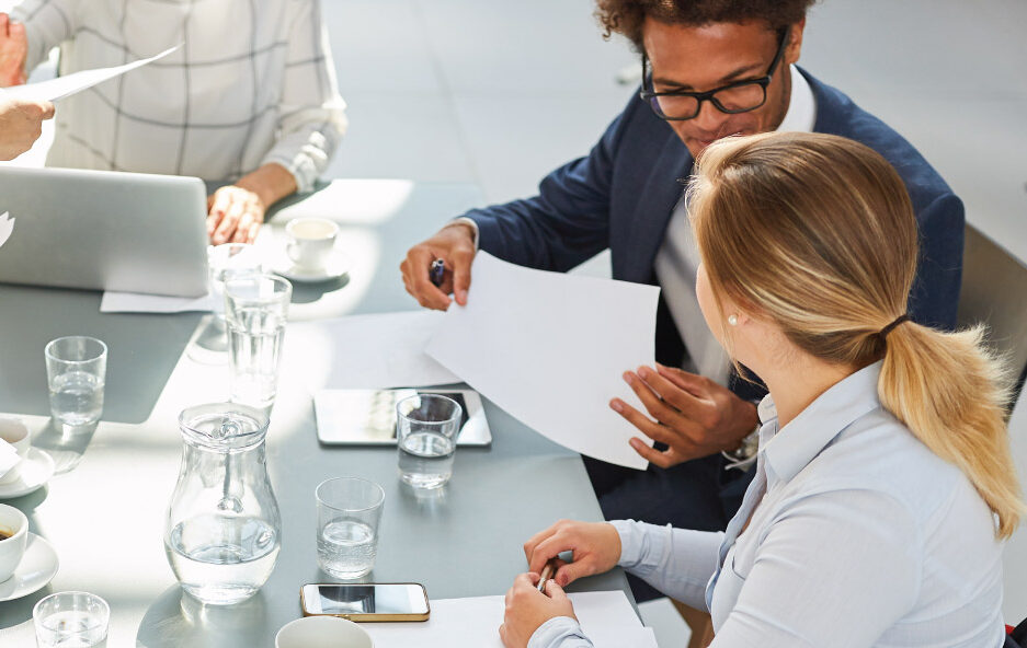 Three people review documents and discuss work at a table with a laptop and notes.