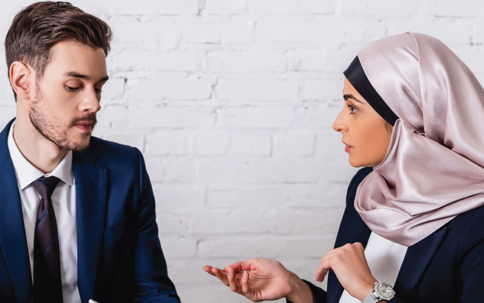 Two people in business attire review documents and talk while seated against a brick wall.