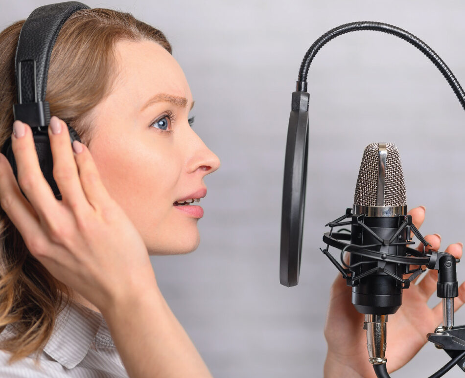 Person wearing headphones speaks into a studio microphone with a pop filter.