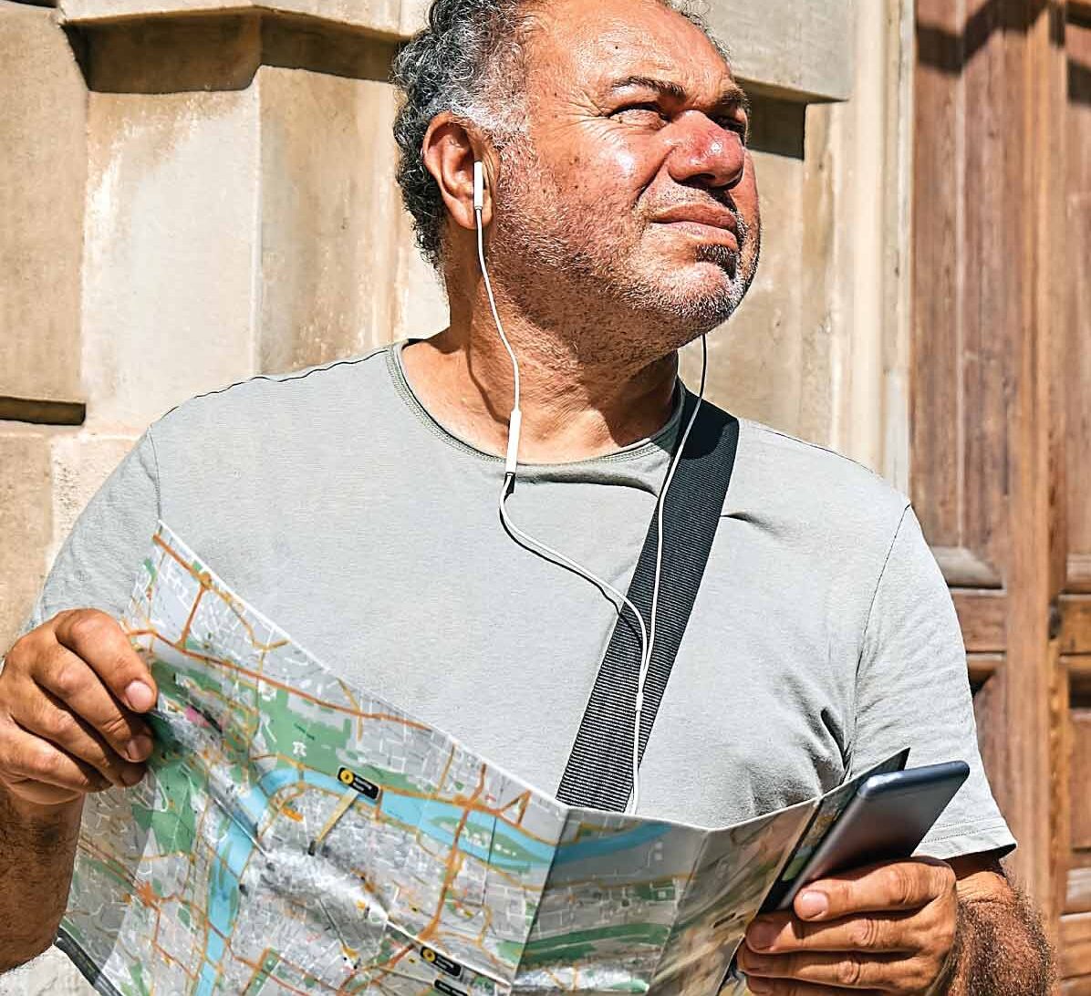 Person holds a paper map and phone while wearing earphones outside a building.