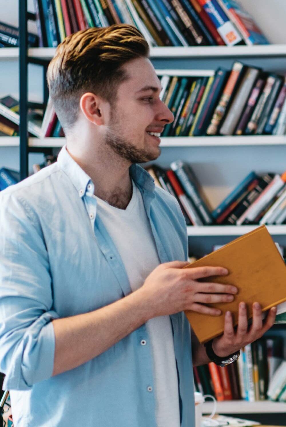Person stands in front of bookshelves holding a book while looking towards the side.