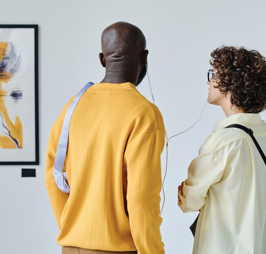 Two people stand side by side using earphones while viewing artwork on a gallery wall.