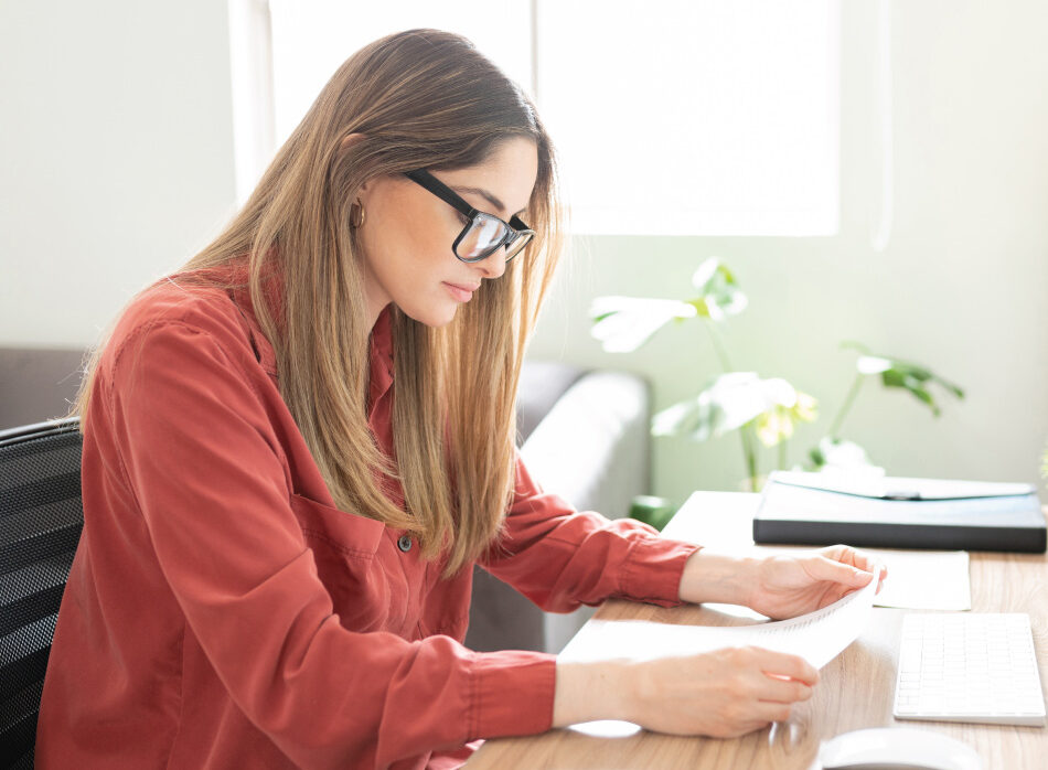 Person wearing glasses reads documents while seated at a desk with a laptop nearby.