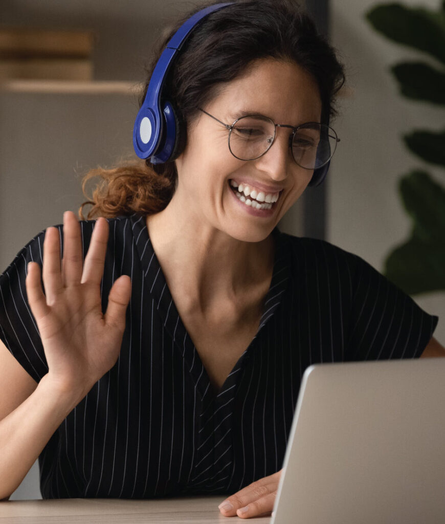 Person wearing headphones waves while using a laptop for a video call at a desk.