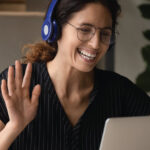 Person wearing headphones waves while using a laptop for a video call at a desk.
