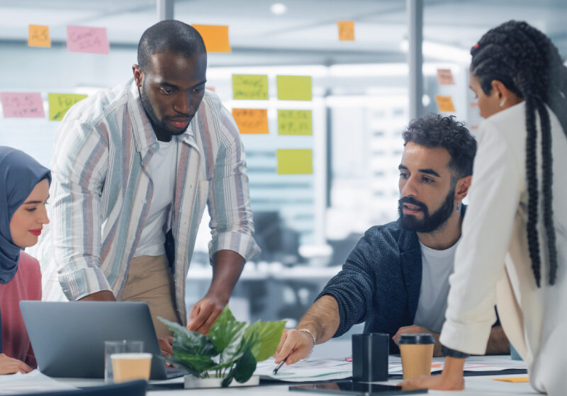 Four people stand around a table reviewing documents and pointing at plans in an office workspace.