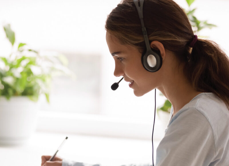 Person wearing a headset writes notes with a pen while seated at a desk indoors.