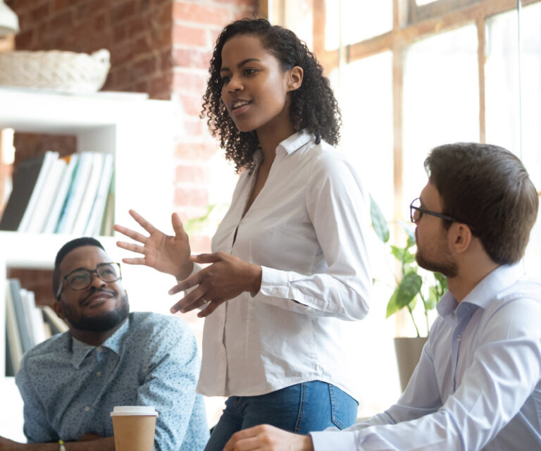 One person stands speaking while two others listen at a table in a bright office space.