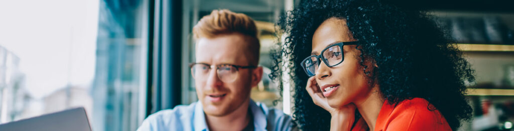 Two people wearing glasses sit close together, looking ahead while working at a desk.